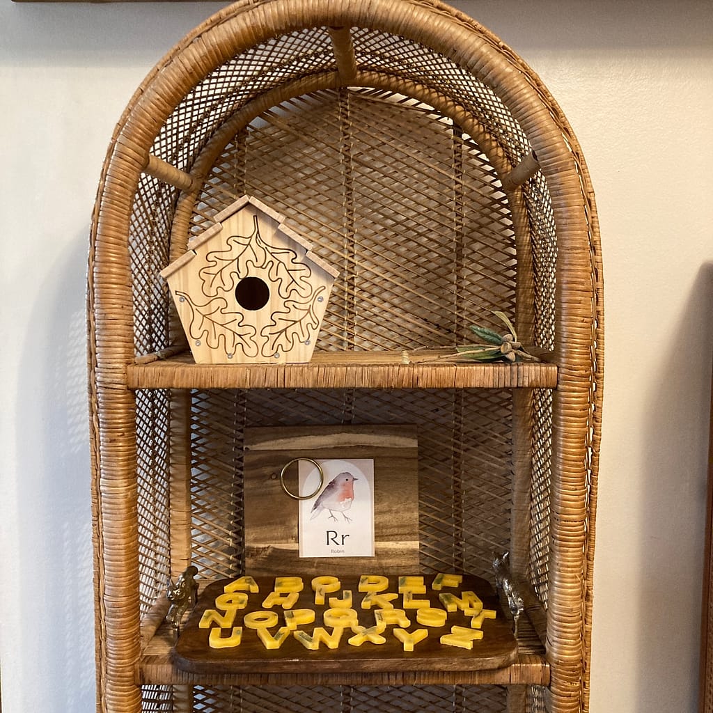 A rattan bookshelf displaying a birdhouse, the beeswax letters, and an easel with a flashcard. 